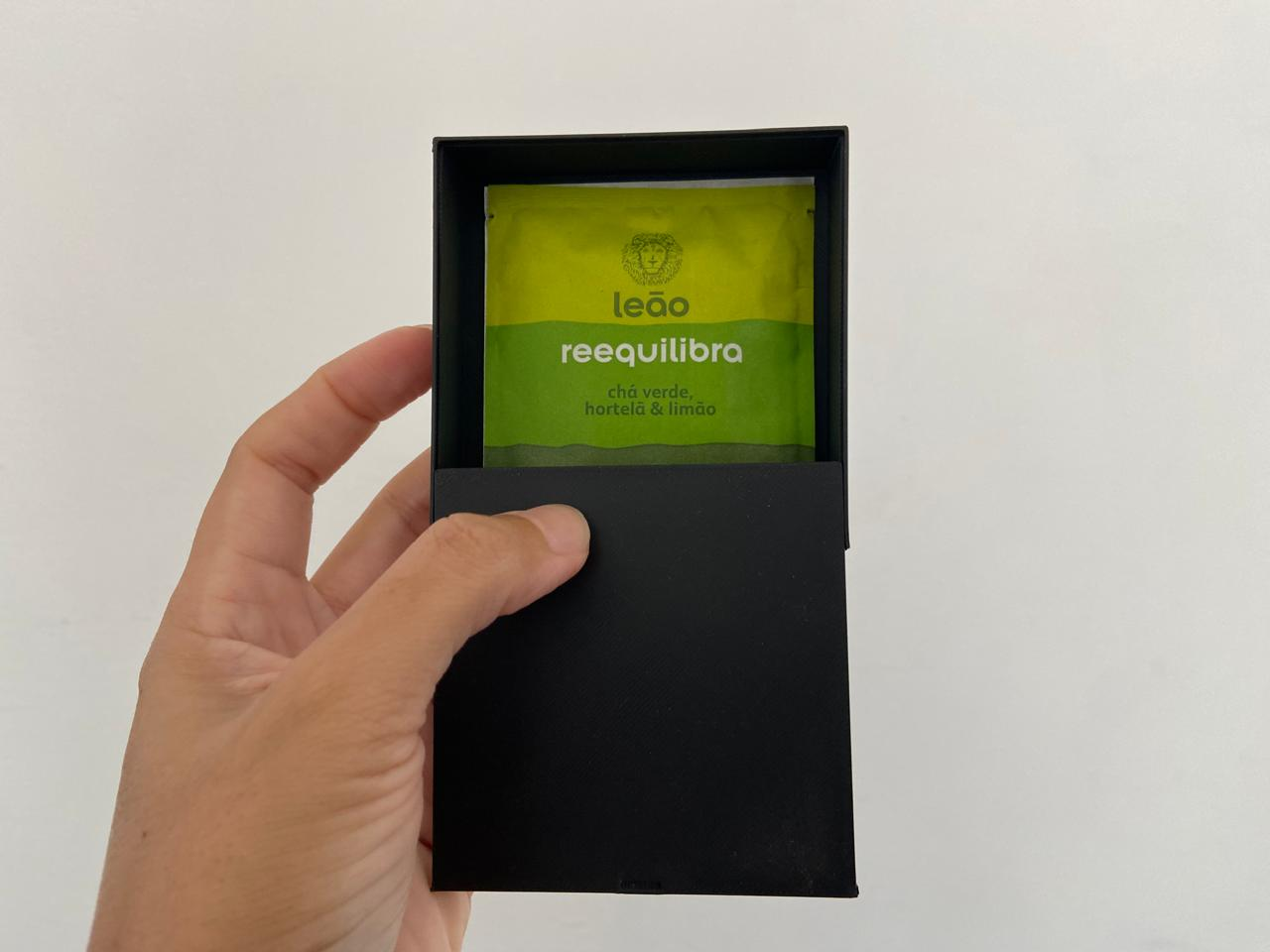 Caixa preta impressa em 3D, aberta e com sachês de chá no interior, sendo segurada na mão.